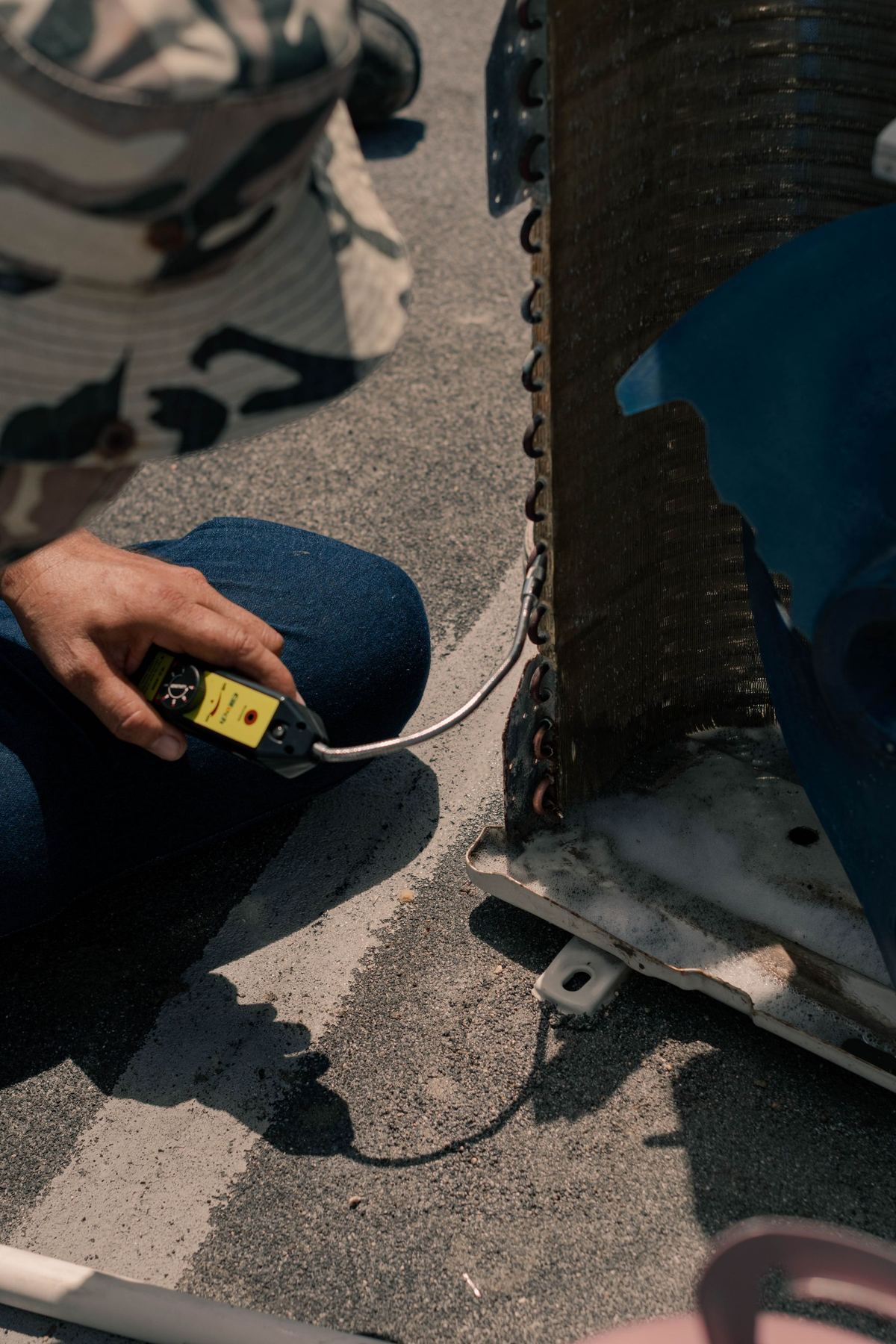 Technician kneeling beside an outdoor air conditioning unit in bright sunlight, checking it with a handheld meter