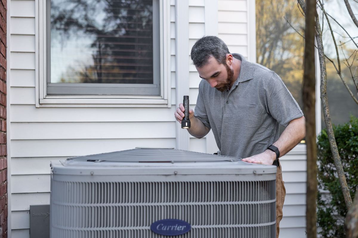 Technician inspecting an outdoor heat pump unit beside a house with a flashlight