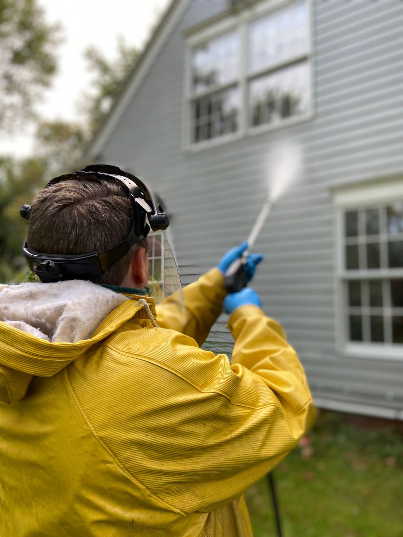 A worker in a yellow rain jacket and face shield washing the light gray siding of a two story house, spray arcing up the wall.